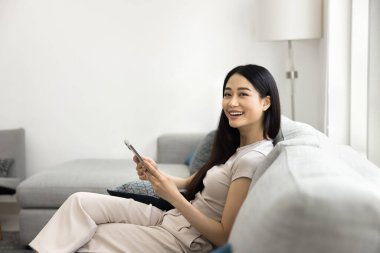 Neşeli genç Asyalı kadın modern koltukta rahat bir şekilde oturuyor, akıllı telefon tutuyor ve kameraya gülümsüyor, ekran zamanının tadını çıkarıyor, hafta sonları evde yeni yapay zeka uygulamaları kullanıyor. Dijital bağlantı, teknoloji