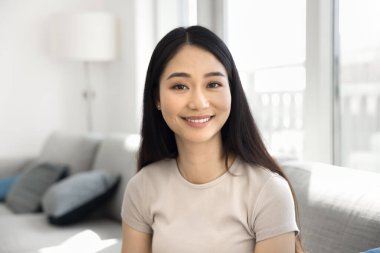 Uzun siyah saçlı güzel bir Asyalı kadın, güneşli oturma odasında kanepede oturmuş kameraya bakıyor, poz veriyor, kendine güven ve pozitiflik yayıyor, hafta sonları evde tek başına dinleniyor, portre çiziyor.