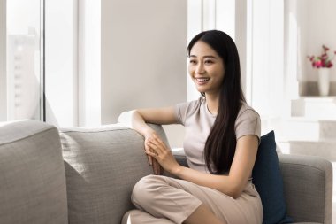 Genç Asyalı kadın, güneşli oturma odasındaki modern kanepede oturuyor, neşeli ve rahat görünüyor, kanepede rahatça dinleniyor, evde huzurlu bir anın tadını çıkarırken gülümsüyor. Dinlenme, kendi evinde rahatlama.