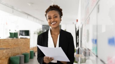 Kendine güvenen genç bir profesyonel. Resmi takım elbiseli mutlu genç iş kadını portresi. Yaratıcı ofis alanında gülümsüyor. Elinde kağıt belgeler, kameraya bakıyor.