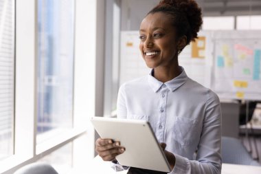 İyimser yaratıcı enerji. Mutlu gülümseyen milenyum iş kadını elinde dijital tabletle ofis penceresinin yanında duruyor fikirleri yansıtıyor başarıyı hırsla hayallere daldırıyor.