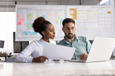 Verimli işbirliği. Çok kültürlü iş adamları ve iki farklı ofis çalışanı birlikte dizüstü bilgisayardaki şirket projesini geliştirmek için dijital görev için basılı raporları analiz ediyorlar.