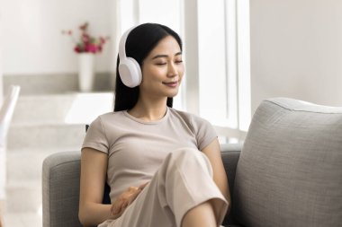 Kapalı gözlü genç Çinli bir kadın oturma odasında koltukta oturuyor, kablosuz kulaklıkla müzik dinliyor, rahat ve huzurlu görünüyor. Hafta sonu hobisi, kaliteli ses, modern teknoloji