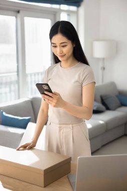 Akıllı telefon standı kullanan Asyalı kadın kutu başında, paket teslimat durumunu takip ediyor, çevrimiçi siparişi onaylıyor, nakliye bildirimini okuyor, alınan öge için gözden geçirme yapıyor, müşteri hizmetleriyle bağlantı kuruyor.