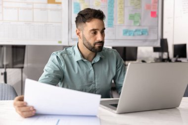 Modern iş akışı. Konsantre genç girişimci strateji pazarlama analisti. Not defterinde çalışıyor. Proje takibi için kağıt ve çevrimiçi kaynaklar kullanarak finansal raporları inceliyor.