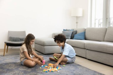 Gülümseyen oğlan ve kız modern oturma odasındaki rahat halıda eğleniyorlar, renkli plastik bloklarla neşe içinde inşa ediyorlar, birlikte odaklanıyorlar ve yaratıcı oyunlarla meşguller. Çocukluk hayal gücü, kardeş bağı.