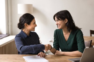 Takım çalışması başarılı. İki gülümseyen genç kadın iş ortağı ofis masasında, proje sözleşmesindeki başarılı ortaklıktan memnun olan defter kağıtlarıyla el sıkışıyor.