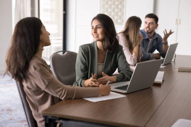 Modern ofisteki dizüstü bilgisayarlarla masada oturan bir grup genç profesyonel, görev veya proje ayrıntılarını tartışarak olumlu ve dostane bir sohbete giren iki bayan meslektaşa odaklanıyor. Takım çalışması.
