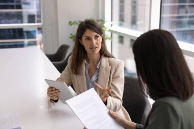 Tablet tutan Kafkasyalı genç bir iş kadını, kadın iş arkadaşına, çalışanına, kağıt belgeli bir müşteriye danışmanlık yapıyor, masadaki iş arkadaşıyla konuşuyor.
