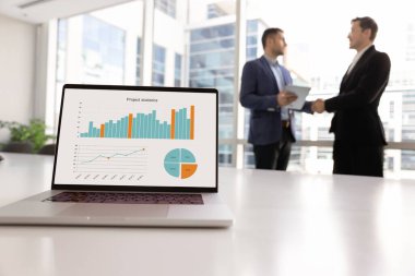 Bilgisayarında büyüme grafikleri olan pazarlama analistlerinin çalışma masası ve arka planda çalışan ve tokalaşan iş arkadaşları. Bilgisayar ekranında ticari bilgi verileri