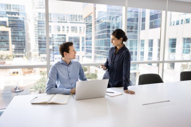 Dostça akıl hocalığı. Gülümseyen milenyum kadın akıl hocası koçu çalışma masasında duruyor ve genç erkek meslektaşı danışman stajyerin modern ofiste defter başında oturması hakkında görüşlerini bildiriyor.