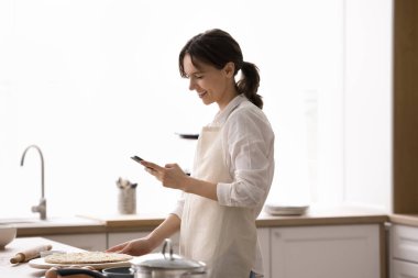 Ev yapımı için dijital yardım. Gülümseyen genç kadın mutfak önlüğüyle tezgahın yanında pizza hamuru düzleştirilmiş halde duruyor. Yemek saati video rehberi, cep telefonu kullanarak internet tarifini takip ediyor.