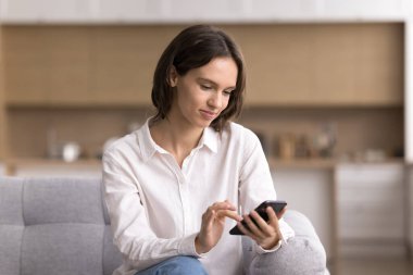 Evdeki dijital yaşam tarzı. Sakin gülümseyen genç kadın oturma odasında akıllı telefonuyla internetten mesajlaşarak sosyal medya alışverişini rahat bir kanepeden yapıyor.