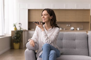 Sesden mesaj teknolojisine. Gülümseyen milenyum kadını, modern akıllı telefon hoparlörünü kullanarak kanepeye oturur sesli mesaj kaydeder, konuşma komutlarını ai app kontrol akıllı ev aygıtlarına verir, ses içeriği oluşturur.