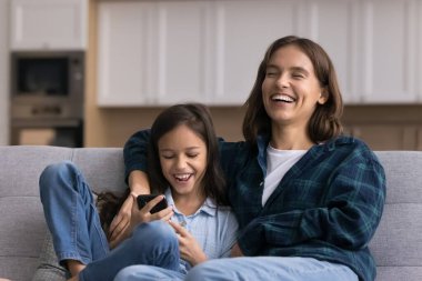 Aletlerle kahkaha. Neşeli bin yıllık anne yetişkin kız kardeş gülüyor küçük kız kardeş cep telefonu kullanıyor evde kanepede eğlenceli vakit geçiriyor sevimli çizgi film klipleri seyrediyor