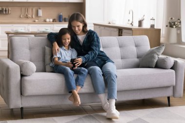 Cihaz ve ebeveyn kontrolü kullanarak. Mutlu küçük kız kucaklaşması... modern stüdyo dairesindeki rahat koltukta sevgi dolu anne dadıya... sosyal medyada dijital anları paylaşarak... telefon üzerinden video izleyerek.