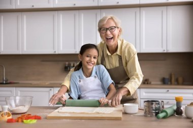 İki nesildir aşçıyım. Portre gülen yaşlı, yetişkin bir büyükanne modern mutfak masasında duruyor. Yemek blogu için ev yapımı fırın için hamur rulosu yapmaktan dikkati dağılmış kameraya bakın.