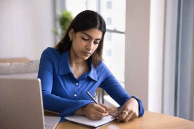 Odaklanmış genç Hintli kadın not defterine notlar yazıyor. Ev işi masasında, dizüstü bilgisayarda oturuyor, günlük plan yapıyor, program yapıyor, bilgisayardan webinar izliyor.