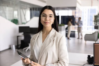 Kendine güvenen genç Asyalı iş adamı gözlüklü, kameraya bakan, açık ofis çalışma alanında duran, dijital masa bilgisayarı tutan, vesikalık portre için poz veren.