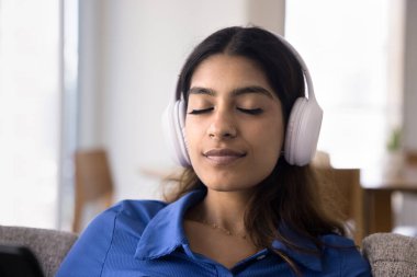 Rahat, huzurlu, büyük kulaklıklar takan genç bir Hintli kadın, meditasyon müziği dinliyor, gözleri kapalı, cep telefonu tutuyor, rahatlamanın tadını çıkarıyor, ses terapisi yapıyor.
