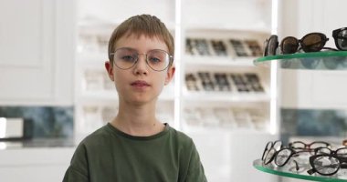 Modern optik dükkanında tek başına duran, yeni gözlüklü mutlu küçük okul çağındaki çocuğun portresi. Göz sağlığı, pediatrik optometri, net görüş ve şık gözlük yoluyla kendine güven