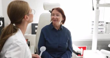 Göz doktoru kliniğinde yaşlı bir kadınla göz muayenesi sırasında konuşan kadın optometrist, hasta dikkatle dinlerken bir sonraki teşhis adımlarını açıklıyor. Modern göz kontrolü hizmetleri