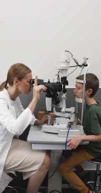 Paltolu kadın optometrist modern klinik ofisinde kesik lamba mikroskobu kullanarak yakışıklı çocuk için göz muayenesi yapıyor. Görme testi, pediatrik göz bakımı, tıbbi hizmetler ve erken görme kontrolü.