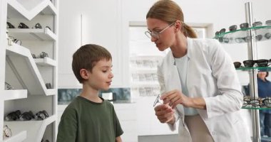 Beyaz önlüklü dost canlısı bayan gözlükçü, modern optik mağazaya uyum sağlarken küçük sevimli çocuğun yeni gözlük takmasına yardım ediyor. Çocuk bakımı, vizyon düzeltmesi, profesyonel optometri hizmetleri