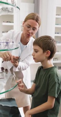 Küçük tatlı çocuğa yardım eden, ceketli neşeli bayan gözlükçü modern optik mağazada ayakta duran güneş gözlüklerini seçiyor, profesyonel göz bakımı, pediatrik optometri hizmeti veriyor. Vizyon ve kişisel stil