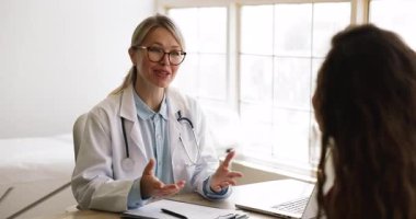 Laboratuvar önlüklü ve gözlüklü çekici bir kadın terapist teşhis veya tedavi planını tartışıyor, reçeteli ilaç açıklamaları yapıyor, klinikteki masada oturan kadın hastayla kurşun konuşmalar yapıyor.