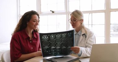 Tıbbi konsültasyon sırasında genç kadın hastaya beyin MR 'ı veya tomografisi gösteren kadın nörolog veya radyoloji uzmanı, modern klinik ofisinde teşhis veya nörolojik tedavi planını tartışıyor.