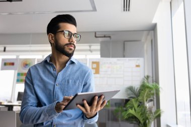 Düşünceli hırslı genç Orta Doğulu profesyonel bir adam tablet üzerinde online iş sürecini yönetiyor, pencerenin yanında duruyor, başka tarafa bakıyor, online pazarlama stratejisi düşünüyor.