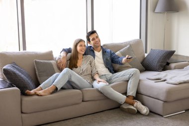 Genç çift oturma odasında rahat bir kanepede dinleniyor, televizyon seyrediyor, uzaktan kumandayla içeriği seçiyor, kanal değiştiriyor, hafta sonu rahatlığının, yayın eğlencesinin ve modern yaşam tarzının tadını çıkarıyor.