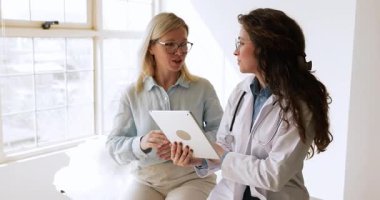 Beyaz önlüklü genç bayan doktor klinik muayene odasında dijital tablet kullanan orta yaşlı bir kadına teşhis veya tedavi seçeneklerini açıklıyor. Sağlık hizmetleri