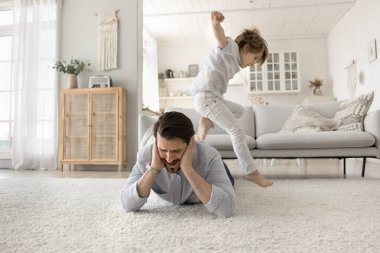 Ebeveynler aşırı yüklendi. Stresli sinirli baba, halıya uzanıp kulaklarını palmiyelerle kapatıyor. Yaramaz, hiperaktif çocuğun gürültüsünden boğulmuş bir şekilde ebeveyn enerjisiyle zıplıyor.