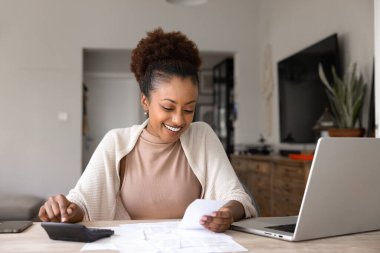 Pozitif genç siyahi kadın evde muhasebe yapıyor, kar sayıyor, tasarruf ediyor, hesap makinesinde ikramiye alıyor, makbuzları analiz ediyor, kağıt faturaları, dizüstü bilgisayarında faturalar, gülümsüyor.