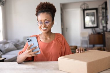 Cep telefonu kullanan, evdeki lojistik karton kutuya dokunan, teslimat servisini sipariş eden, paket gönderen, cep telefonuyla yazan güzel ve genç bir Afrikalı kadın.