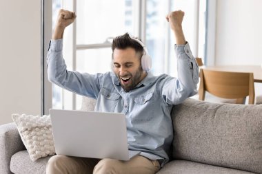 Kablosuz kulaklıklı mutlu genç adam kanepeye dizüstü bilgisayarla oturur, zafer ve heyecan dolu bir jest ile kollarını kaldırır, zaferi kutlar, online oyun kazanır, favori futbol takımının evdeki başarısının tadını çıkarır.