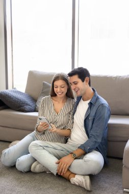 Genç çift oturma odasında akıllı telefon kullanıyor, video konferansına katılıyor, banka veya yatırım uygulamalarını kontrol ediyor, internetten video izliyor, bilet ya da rezervasyon yaptırıyor, fotoğraf albümüne kayıyor