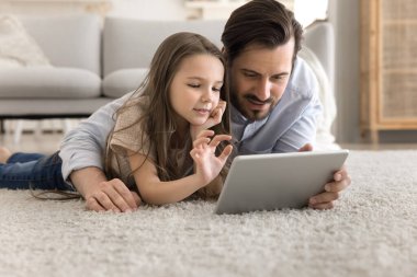 Çevrimiçi eğitim aktivitesi. İlgili babanın portresi sıcak halının üzerinde yatarken küçük kız çocuğuna sarılıp dijital tablet üzerindeki uygulama çalışmasını keşfetmede yardımcı oluyor.