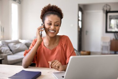 Uzaktan çalışma için dijital teknolojiyi kullanan mutlu genç siyahi iş kadını, ev işyerinde oturuyor, cep telefonuyla konuşuyor, gülümsüyor, gülüyor, dizüstü bilgisayarda yazıyor.