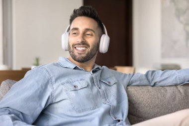 Kablosuz kulaklıklı İspanyol bir adam, içeride koltukta dinleniyor, mutlu bir gülümsemeyle yan yana bakıyor, popüler ya da enstrümantal müzik dinliyor, hafta sonları podcast veya evde sesli kitapların keyfini çıkarıyor. Hobi, teknoloji