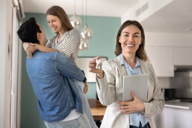 Kameraya ev anahtarlarıyla poz veren mutlu kadın emlakçı arka planda kucaklaşan çift taşınma gününü ilk emlak alımını kutluyor. Başarılı ev sahipliği, hayaller gerçek oluyor, emlak hizmetleri.