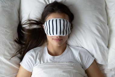 Kaliteli gece uykusu. Yumuşak battaniyenin altında, rahat bir yatakta uyuyan huzurlu, sakin, genç bir bayan. Uyku maskesiyle kaplı gözleri, rahat bir rüya için günlük kendi kendine bakım rutininin tadını çıkarıyor. Yukarıdan üst görünüm