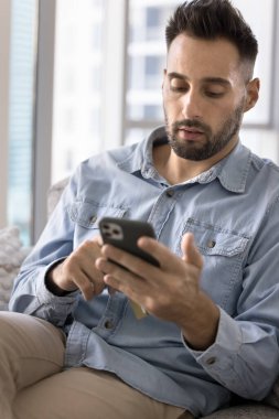 Odaklanmış İspanyol adam evdeki koltukta oturuyor, akıllı telefon kullanıyor, arkadaşlarıyla sohbet ediyor, sosyal medyada geziniyor, randevu veya hizmet alıyor, cep telefonu uygulaması üzerinden mesaj veya bildirimle cevap veriyor. Teknoloji