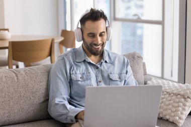 Modern oturma odasındaki kanepede rahatça otururken dizüstü bilgisayar ve kablosuz kulaklık kullanarak İspanyol gömlekli gülümseyen genç adam. Uzak çalışma, dijital iletişim veya çevrimiçi öğrenme etkinliği