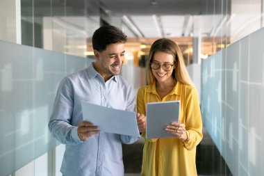 Satış raporlarını karşılaştıran, tablet üzerinde kağıt ve elektronik içeriği analiz eden, ofis koridorunda duran, konuşan, finansal analitik verileri tartışan mutlu genç iş arkadaşları