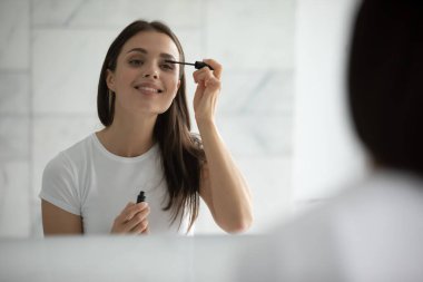 Sabah kozmetik ayini. Beyaz tişörtlü güzel, gülümseyen genç bir kadın doğal güzelliği vurgulamak için basit göz makyajı, makyaj kirpikleri ve siyah rimel sür. Aynada kafasından vurulmuş.