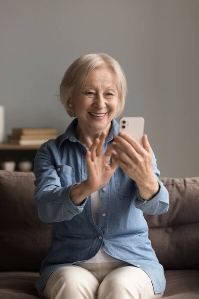 Tech savvy aging. Happy confident older lady sit on cozy sofa enjoy mobile phone app with smile create profile picture for social media edit selfie photo using face filters watch funny videos on blogs