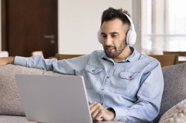 Kulaklıklı İspanyol bir adam dizüstü bilgisayardan online içeriği izliyor, internette boş zaman geçiriyor, dijital abonelik hizmetlerini kullanarak film izliyor, sanal toplantıya ya da video çağrısına katılıyor.